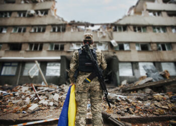 Ukrainian military woman with the Ukrainian flag in her hands on the background of an exploded house