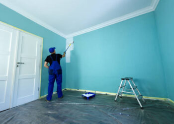 Man in a working overall is painting the wall in white color